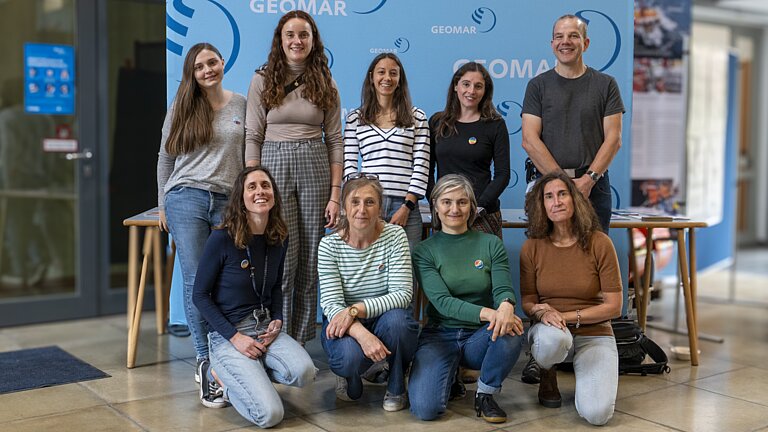 Neun Personen posieren für die Kamera. Es sind acht Frauen und ein Mann. Fünf stehen, vier knieen davor