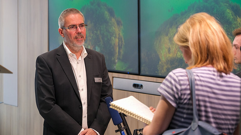 A man stands in front of a screen showing a picture of a diver under water. A woman and a man with a notepad and microphone are interviewing him.