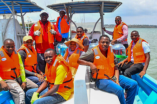 Zehn dunkelhäutige Personen mit lauchtend orangen Schwimmwesten auf einem blauen Boot