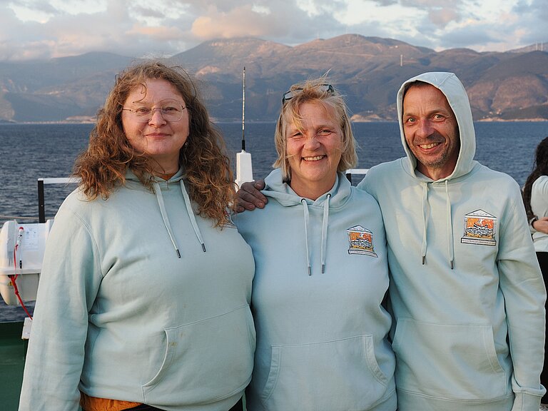 Zwei Frauen und ein Mann stehen an Bord eines Schiffes vor der Silhouette einer felsigen Küste