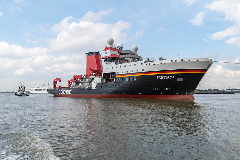A large ship with a black and red hull and white superstructure is being towed across the water on a towline.