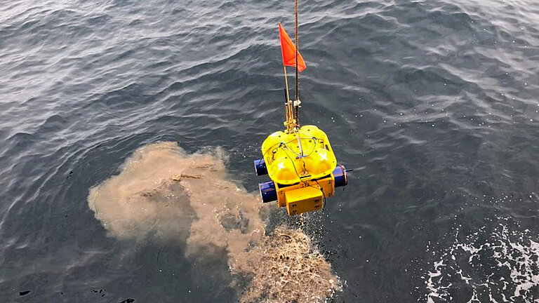 Ein gelbes kastenförmiges Messgerät mit einem roten Fähnchen wird vom Meeresboden wieder an die Wasseroberfläche gehievt, eine sandfarbene Sedimentwolke im Wasser hinterlassend