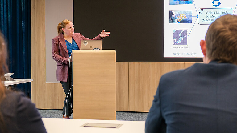 Eine Frau an einem Rednerpult zeigt auf eine an die Wand projizierte Präsentation