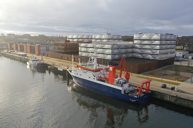 Luftaufnahme eines Forschungszentrums an einem Flussufer, an der Pier liegen ein kleineres und ein größeres Forschungsschiff; Morgenstimmung