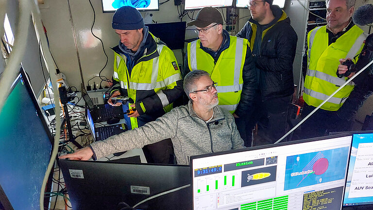 Ein Mann mit grauem Haar und Bart sitzt an einem Schreibtisch mit vielen Monitoren und zeigt auf einen Monitor, auf dem ein Unterwasserbild zu sehen ist, vier Männer, teils mit gelben Warnwesten, schauen ihm über die Schulter.