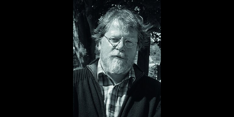 Schwarz-weiß-Foto eines älteren Mannes mit runder Brille, wuscheligem Haar und Vollbart. Er trägt ein kariertes Hemd unter einem wollenen Pullover mit Reißverschluss.