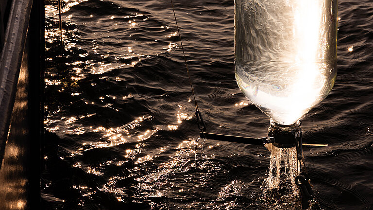Ein gläserner Behälter wird, mit Wasser gefüllt, aus dem Meer gezogen. Die Morgensonne scheint durch das Glas.