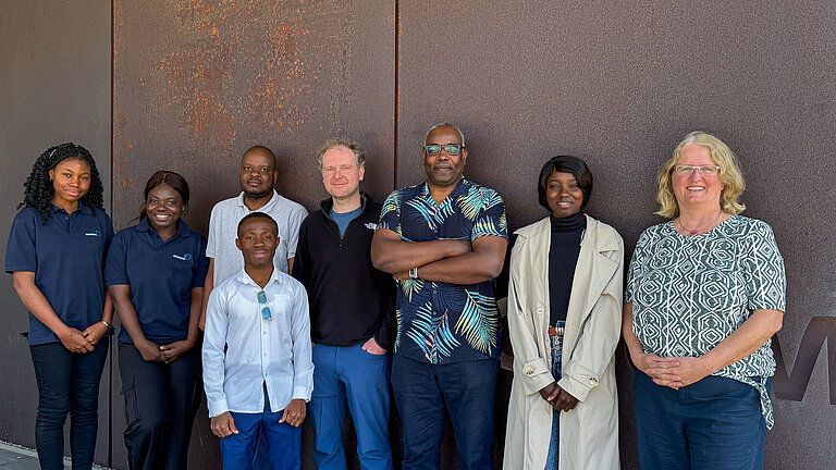 Acht Personen stehen vor einer rostfarbenen Wand