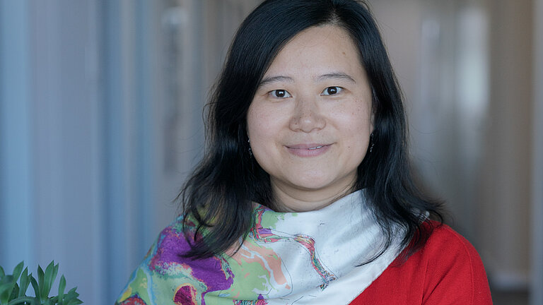 [Translate to English:] Eine Frau mit asiatischen Gesichtszügen und schwarzen halblangen Haaren lehnt an einem Stehtisch und lächelt in die Kamera. Sie trägt einen roten Pullover und ein weißes Halstuch mit Blumen.