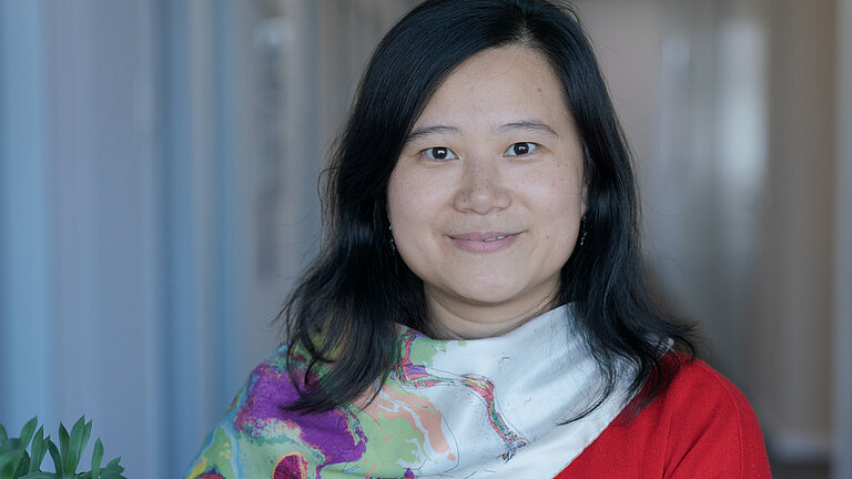 Eine Frau mit asiatischen Gesichtszügen und schwarzen halblangen Haaren lehnt an einem Stehtisch und lächelt in die Kamera. Sie trägt einen roten Pullover und ein weißes Halstuch mit Blumen.