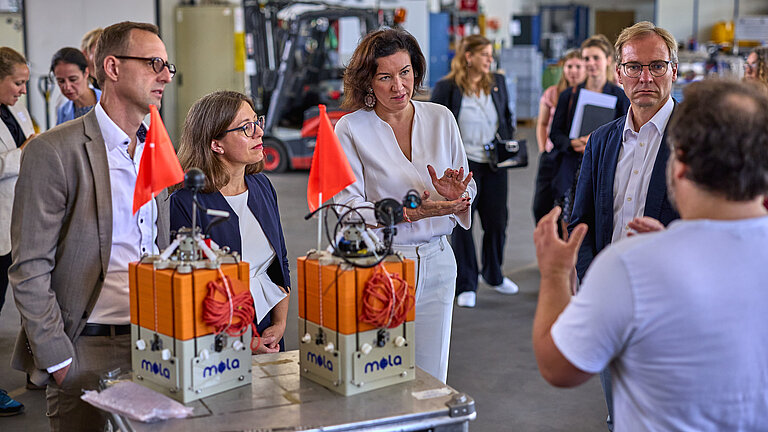 Eine Gruppe formell gekleideter Personen steht im Halbkreis und hört einem Wissenschaftler im weißen T-Shirt zu, im Vordergrund stehen kleine orangefarbene Messgeräte