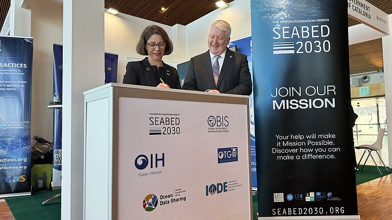 Eine Frau und ein Mann an einem Messestand lächeln und unterschreiben ein Dokument