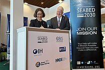 Eine Frau und ein Mann an einem Messestand lächeln und unterschreiben ein Dokument