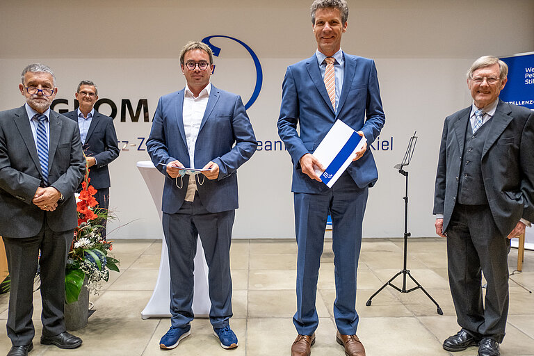 Fünf Männer stehen mit Abstand zueeinander im GEOMAR-Hörsaal. Die beiden in der Mitte halten Urkunden in der Hand. 