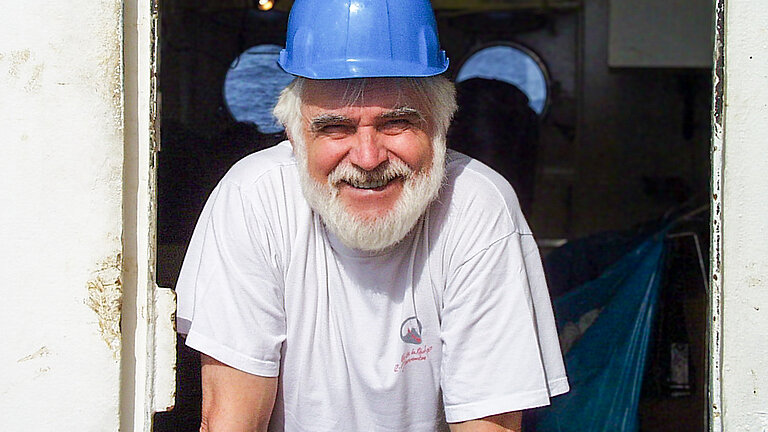 Ein älterer Mann mit weißen Haaren und weißem Vollbart mit einem blauen Sicherheitshelm auf einem Schiff