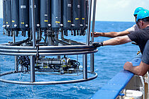 Mitarbeiter auf einen Schiff mit blauen Helmen greifen nach dem Stahlrahmen eines Kranzwasserschöpfers, der aus dem Wasser gehievt wird.