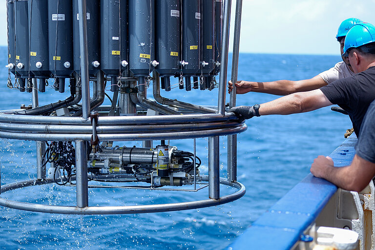 Mitarbeiter auf einen Schiff mit blauen Helmen greifen nach dem Stahlrahmen eines Kranzwasserschöpfers, der aus dem Wasser gehievt wird.