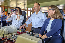 Ein Schiffskapitän auf der Brücke mit zwei Frauen in Hosenanzügen neben sich, eine schaut durch ein Fernglas