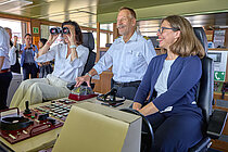 A ship's captain on the bridge with two women in trouser suits beside him, one looking through binoculars.