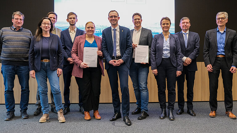 Eine Gruppe Menschen steht vor einer großen Projektionsfläche und schaut in die Kamera