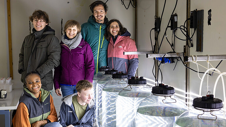 Eine Gruppe von fünf jungen Forschenden schart sich glücklich um die mit Meerwasser befüllten Behälter im Labor.
