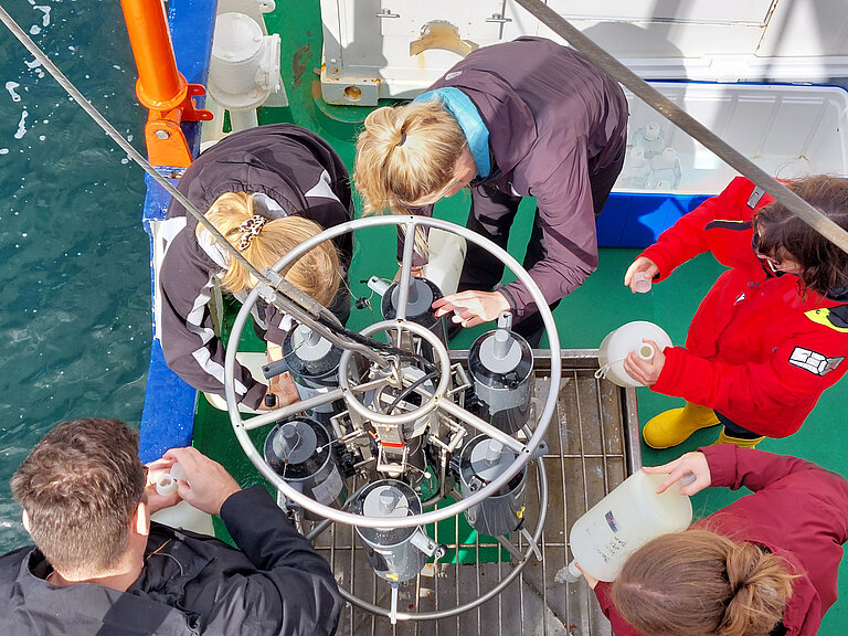 Junge Forschende stehen um einen Kranzwasserschöpfer an Bord eines Schiffes und füllen Wasserproben ab