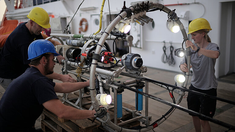 Drei Personen arbeiten an einem Stahlreifen, der mit Scheinwerfern und Kameras ausgestattet ist an Bord eines Schiffes