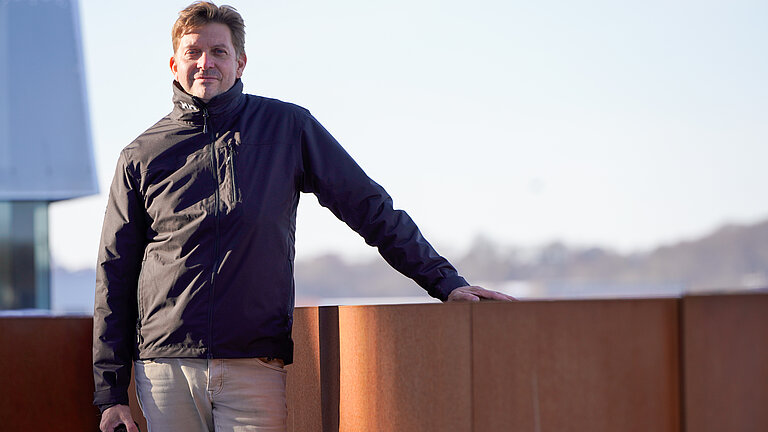 Ein Mann steht an eine rostroten Balustrade, im Hintergrund in der Ferne die Silhouette einer Stadt