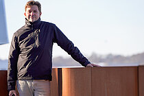 Ein Mann steht an eine rostroten Balustrade, im Hintergrund in der Ferne die Silhouette einer Stadt