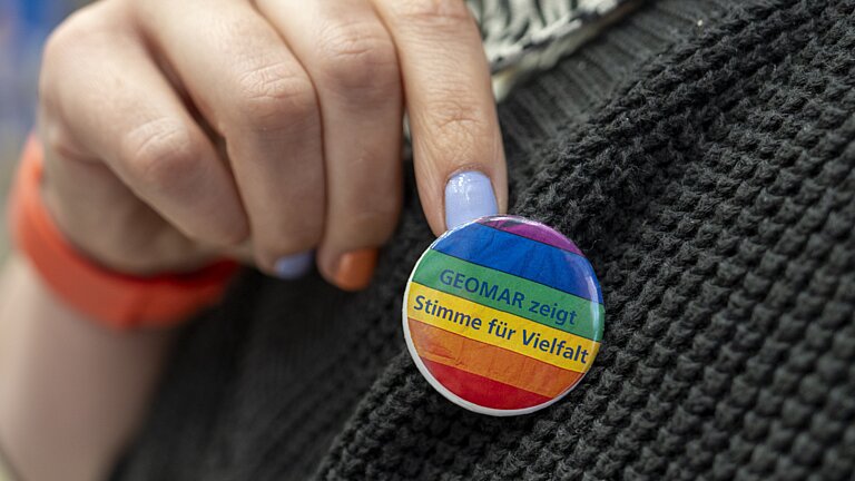 Eine Hand mit hellblauem Nagellack zeigt einen Button mit Regenbogenfarben. Der Button ist an der Jacke befestigt. Gesicht undKörper der Person befinden sich außerhalb des Bildes.