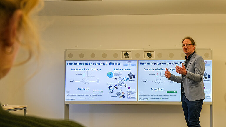 Ein Mann steht vor einer digitalen Tafel, im Vordergrund ist die Silhouette einer Frau zu sehen. 