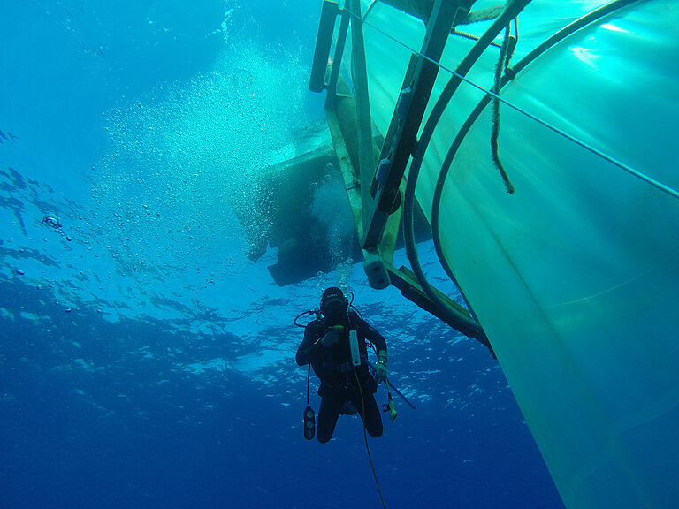 Ein Taucher im blau scheinenden Wasser schwimmt an einem großen farblosen Tank aus Plastikfolie vorbei