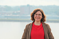 Eine junge Frau steht vor einer Wasserfläche, im Hintergrund Hafenanlagen