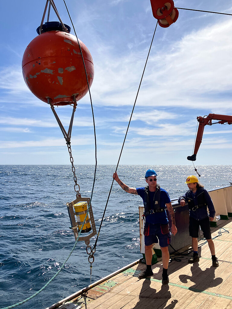 Zwei Personen stehen an Deck eines Schiffs und bringen eine Verankerung aus. 