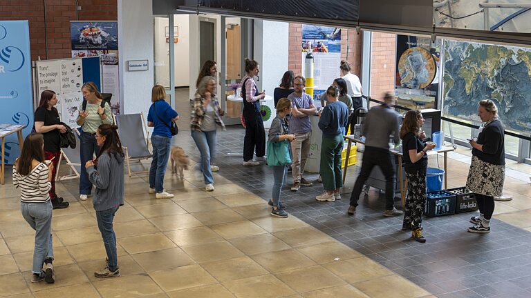 Mehrere Menschen sind in einem großen Raum. Sie stehen in kleinen Gruppen und unterhalten sich.