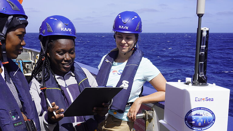Drei Personen tragen Helme und stehen auf einem Forschungsschiff