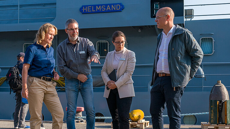 Vier Personen stehen auf einer Pier vor einem Marineschiff