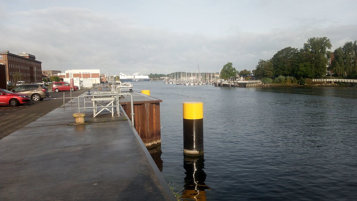 Man sieht den Kai vor dem Geomar-Gebäude und den Fluss Schwntine. Aus dem Wasser ragen zwei große schwarze Säulen, die den Bereich markieren und schützen, an dem die Pipeline das Institut erreicht.