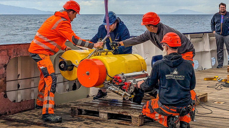 Männer mit Schutzhelmen an Bord eines Forschungsschiffes bringen ein großes Messgerät in Position, das ins Wasser gelassen werden soll