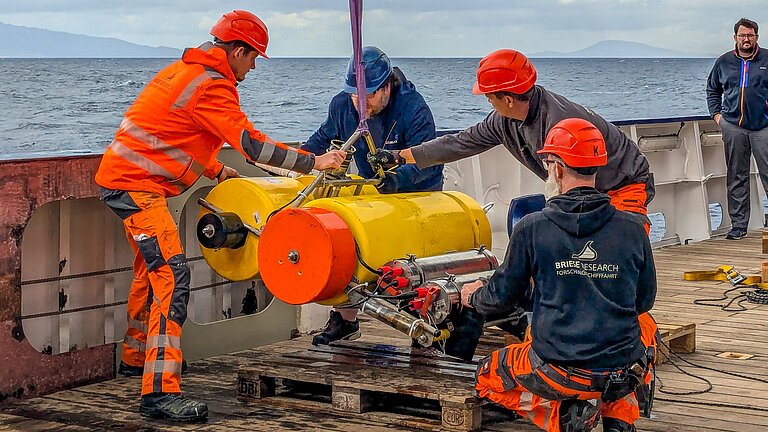 Men wearing protective helmets on board a research vessel position a large measuring device that is to be lowered into the water.