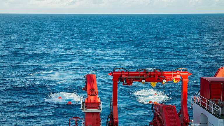 Blue sea, the red crane of a research vessel can be seen at the bottom right of the picture, two red buoys are being towed behind the ship