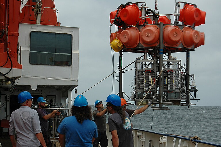 Man sieht die Reling des Schiffs, über die gerade mit einem Schiffskran ein großes Gestell mit orangenen Kugeln und dem gelben Viator-Fahrzeug ins Wasser gelassen wird. Menschen beaufsichtigen den Prozess.