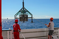Zwei Arbeiter mit roten Schutzhelmen stehen an Bord eines Forschungsschiffes, während ein Forschungsgerät an Bord gehievt wird 