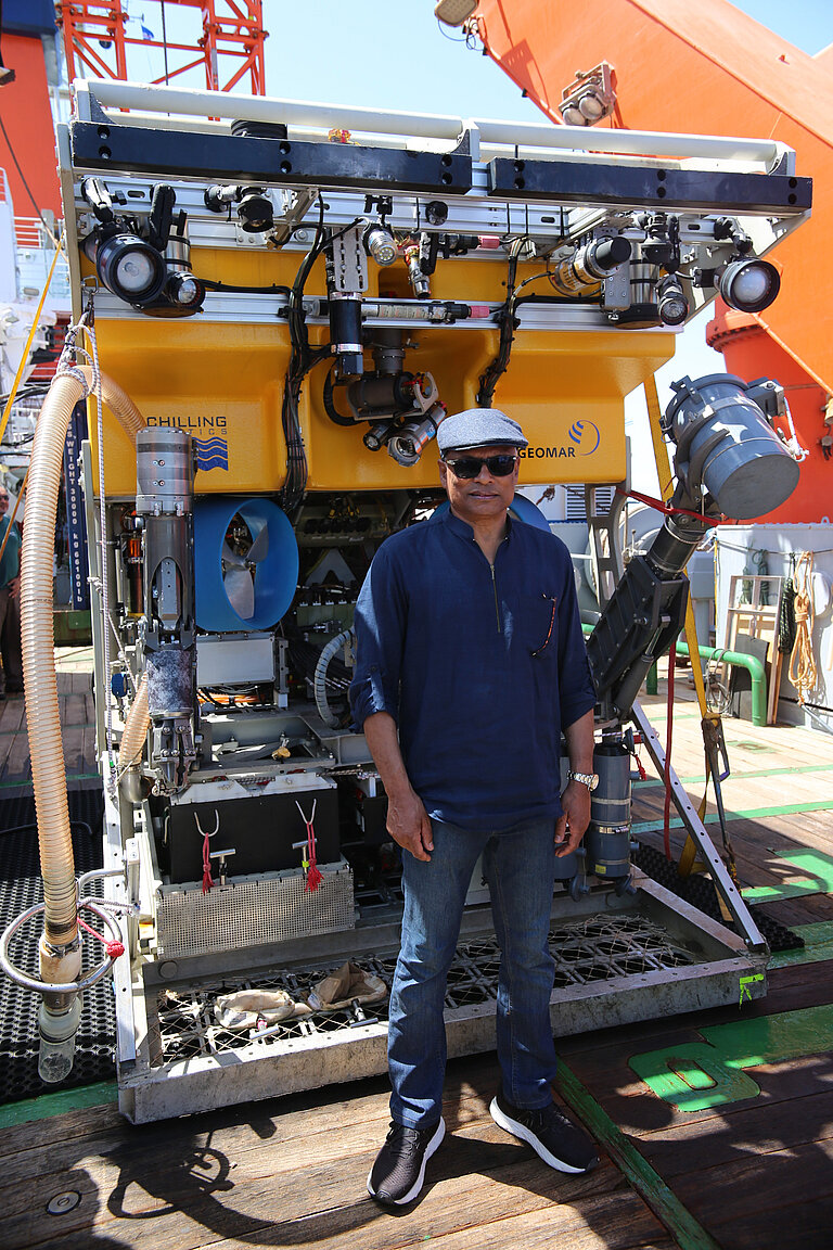 Ein dunkelhäutiger M;ann in Jeans, blauem T-Shirt und mit Schiebermütze und Sonnenbrille posiert vor einem gelben Tauchroboter an Bord eines Schiffes