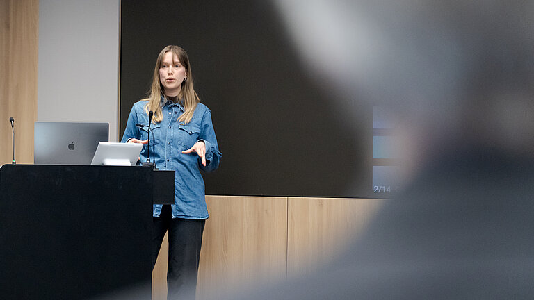 Eine junge Frau mit langen blonden Haaren und einem Jeanshemd steht an einem Pult und spricht zu Publikum.