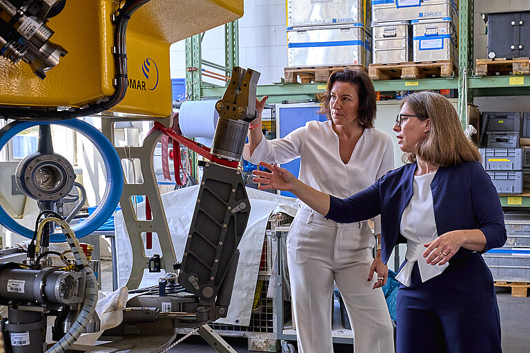 Two women in elegant trouser suits look at an underwater robot.