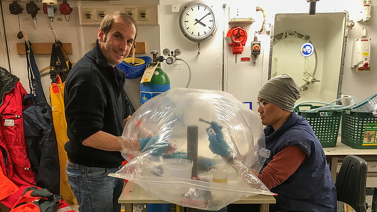 Two men are working in a laboratory on a ship on a sediment core protected by a plastic cover.