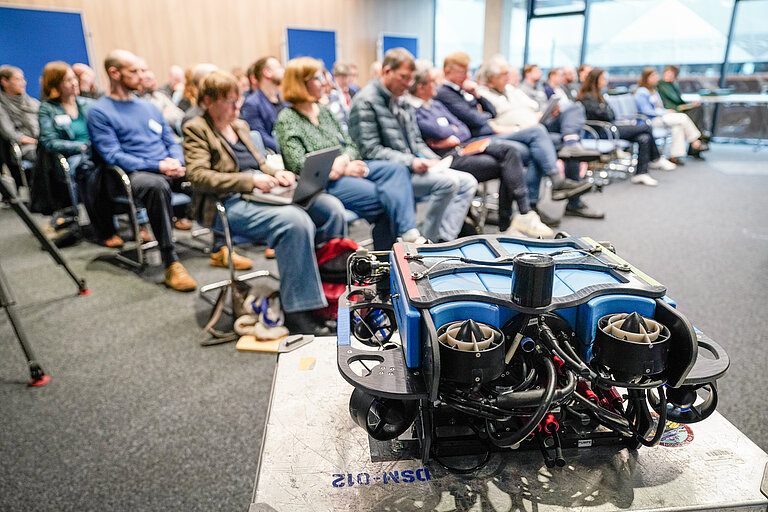 Im Vordergrund ein meereswissenschaftliches Grät, unscharf dahinter ein Konferenzraum, mit Publikum gefüllt