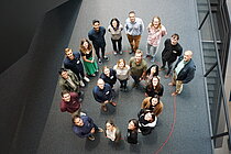 Mehrere Personen stehen auf einem dunkeln Teppich in einer Spirale angeordnet. Sie schauen nach oben zur Kamera. 