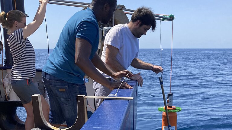Eine Frau und zwei Männer lassen gemeinsam ein Messgerät über die Reling eines Schiffes zu Wasser